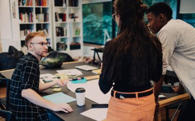 Giyim kuşamında genç meslektaşlar masada çalışma fikirlerini tartışıyor ofisteki bir fincan kahve belgeleriyle.