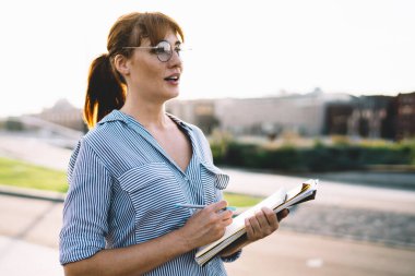Gözlüklü, günlük kıyafetleri olan çekici beyaz kadın güneşli bir günde şehir ortamında geziniyor, güzel hippi kız elinde kitap tutuyor ve hafta sonları güneşli bir günde parkta yürüyor.