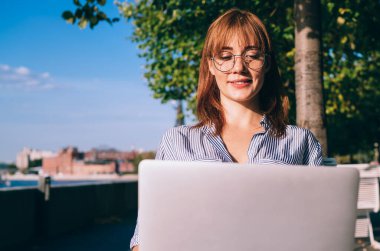 Neşeli kızıl saçlı kadın görüş koruması için serbest çalışıyor video seyrediyor ve laptopta haber okuyor güneşli bir günde dışarıda oturuyor, kadın dijital göçebe şehirde uzaktan çalışıyor.