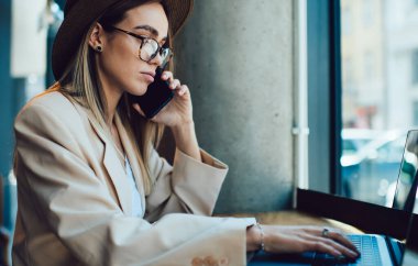 Moda ceketli ve şapkalı ciddi anlamda çekici bir kadın serbest yazar, kafede dizüstü bilgisayarla çalışırken telefonla konuşuyor.