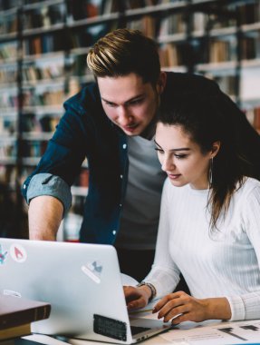 Kot ceketli bir erkek üniversite projesinde çalışan dizüstü bilgisayarı gösteriyor ve kütüphanede masasında oturan bir kadın var.
