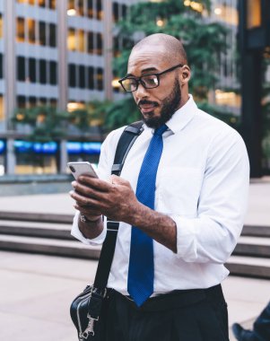 Ciddi ve odaklanmış gözlüklü siyah bir çalışan. Parlayan pencereli binaların yanında dururken patrondan gelen mesajı dikkatle okuyor.