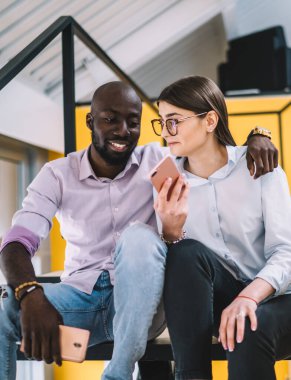 Genç, kel, mutlu bir adam merdivenlerde akıllı telefonlarla konuşurken ve otururken elini kadının omzuna koyuyor.