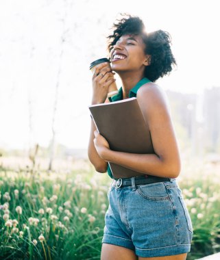 Duygusal mutlu Afrikalı Amerikalı öğrenci elinde kitapla gülüyor kaygısız, koyu tenli, heyecanlı kadın kahveyle güneşli yaz günlerinde parkta eğlenmeye gidiyor.
