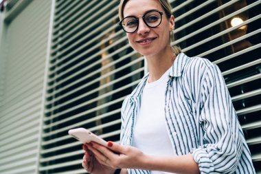 Gözlüklü, rahat giyinen, kameraya bakıp gülümseyen, pencereye yaslanan ve elinde akıllı telefon tutan iyimser bir kadın.