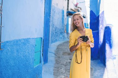 Fas 'ın güneşli bir gününde yaşlı bir kasabanın dar sokağında dikilirken kamerayla gülümseyen ve yüzünü çeviren mutlu kadın turist.