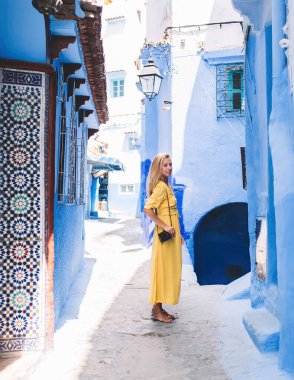 Sarı elbiseli, omzunda fotoğraf makinesi olan genç bir kadın dar bir sokakta mavi duvarlı eski taş binalarla Fas 'ı gezerken görülüyor.