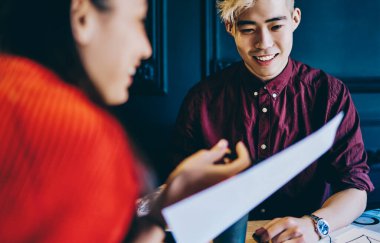 Gündelik giysiler içinde gülümseyen çok ırklı iş arkadaşları ellerinde kağıtlarla bürodaki diyagramlarla masada oturuyorlar.