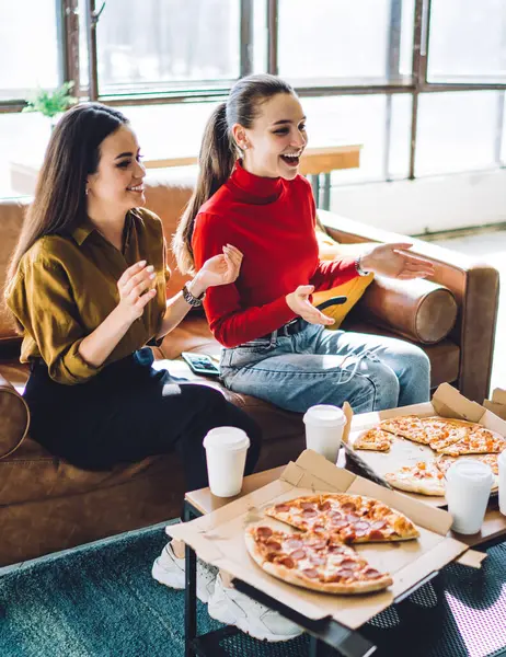 Cheerful people laughing and gesturing while sitting on sofa by table with takeaway cups and delivered pizza at home in daylight