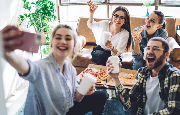 Group of delighted young people with pizza and beverages to go smiling and taking selfie while sitting near table during gathering at home