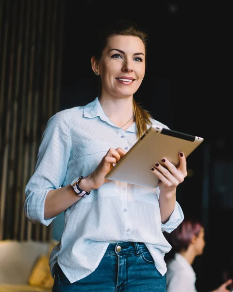 Pleased female manager in casual clothes standing and using tablet while looking away on blurred background with cafe interior and guests