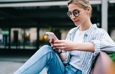 Yuvarlak gözlüklü, modern akıllı telefon kullanan, yeşil çalıların yanında otururken gülümseyen, sıradan giyinen, çekici sarışın kadının yan görüntüsü.