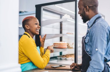 Sarı tişörtlü ve turkuaz önlüklü şık siyahi kadınla konuşurken üniformalı Afrikalı Amerikalı bir adamın gülüşünün yan görüntüsü.