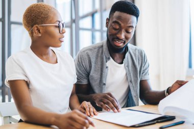 Odaklanmış siyah çift, emlakçının önündeki masada oturmuş dikkatle emlak sözleşmelerini okuyor ve konsantrasyonla konuşuyorlar.