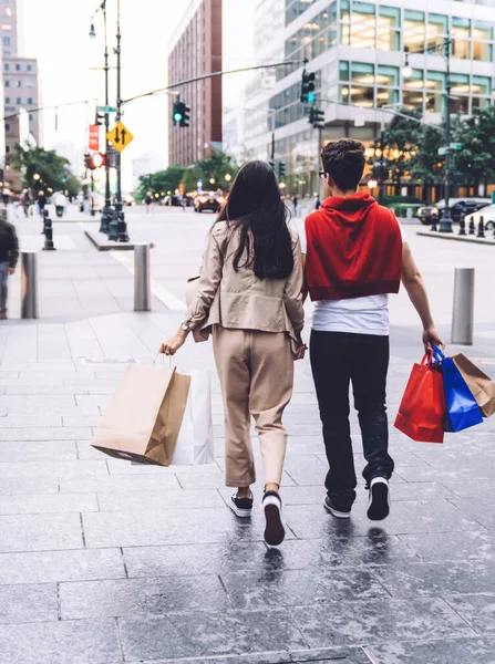 Back view of stylish man and woman holding hands walking on city street with shopping bags against modern city background