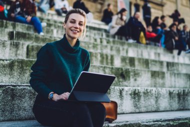 Tablet üzerinde çalışan ve eski üniversite binasının kalabalık merdivenlerinde otururken kameraya bakan mutlu, güler yüzlü kadın serbest yazar.