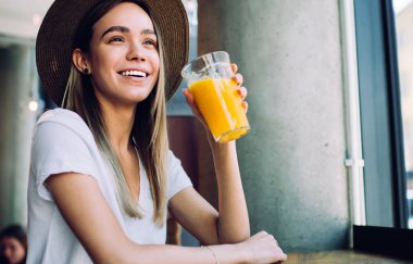Güzel şapkalı mutlu bir bayan pencerenin yanındaki masada oturmuş kafeteryada taze meyve suyunun tadını çıkarırken gülümsüyor ve başka tarafa bakıyor.