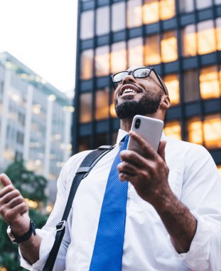 Heyecanlı Afro-Amerikan kel sakallı iş adamı gözlüklü ve akıllı telefonuyla modern ofis binasının önünde başarıyı kutluyor.
