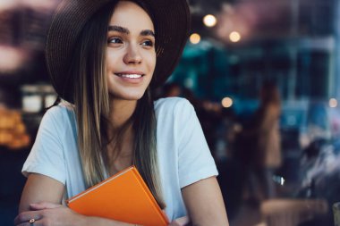 Çekici ve iyimser bayan öğrenci kafenin önünde oturuyor ve turuncu kitabı kucaklarken gülümsüyor ve başka tarafa bakıyor.