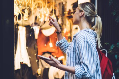 Çizgili tişörtlü, kırmızı sırt çantalı ve vitrindeki kıyafetleri gösteren sarışın kadın turistin yan görüntüsü.