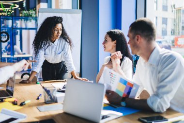 Resmi giysiler içinde kendine güvenen Afrikalı Amerikalı bir kadın ofis binasında çeşitli aletlerle masanın yanında dururken konferans sırasında iş arkadaşlarıyla konuşuyor.