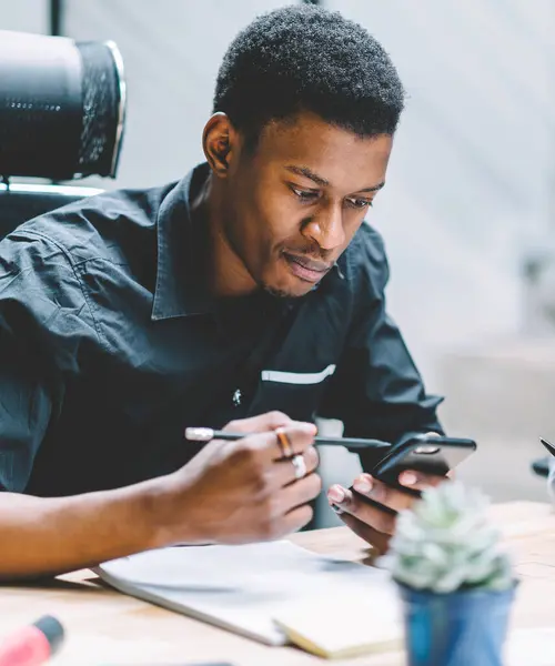 African American male employee searching text on web page with information on cellphone while sitting at desktop with colleague in workspace. Dark skinned young men using mobile phone in office