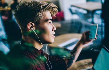 Düşünceli uzaktan kumandalı, kulaklıklı, müzik dinleyen, iş yerinde işe başlamak için dizüstü bilgisayar kullanan düşünceli bir işçinin yan görüntüsü.