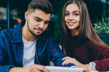 Pozitif genç kadın kamera bakarken erkek arkadaşı taşınabilir pc üzerinden online oyun oynamaya konsantre, erkek ve kadın hipsters video izlemek için cihaz kullanarak kafe terasında oturan