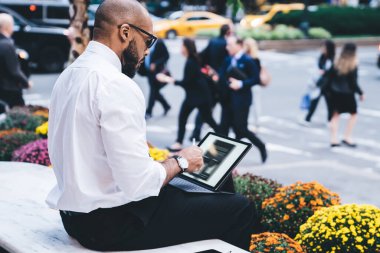 Beyaz tişörtlü, siyah pantolonlu, klavyeli, çiçekli yatağın yanındaki bankta gezen Afro-Amerikan kel adamın yan görüntüsü.