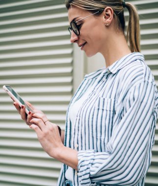 Beyaz tişörtlü, çizgili tişörtlü, gözlüklü, kırmızı tırnaklı, akıllı telefondan kapalı jaluzinin yanındaki videoyu izleyen şık bir kadının yan görüntüsü.