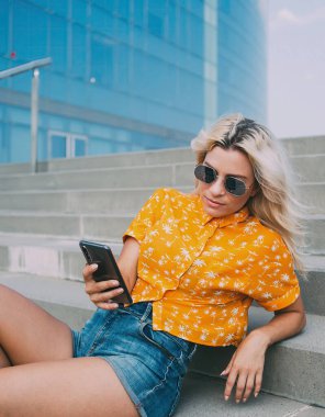 Modern şehrin sokaklarında dinlenirken şık giyinmiş ve güneş gözlüklü rahat genç bayan merdivenlerde uzanıp dinleniyor.