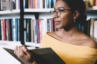 Üniversite kütüphanesindeki akademik araştırmalar sırasında kitaplığa yaslanarak fen dersi okuyan düşünceli Afrikalı Amerikalı kadın gözlerini kaçırıyor.