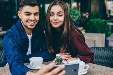 Kafe terasında masada oturan cep telefonu kamerası selfie için poz neşeli erkek ve kadın genç hipsters, kahve ile tarih boyunca cep telefonu üzerinden fotoğraf yapma çift birlikte zaman keyfini çıkarın