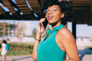 Moda gözlüklü mutlu genç bir kadın yazın açık havada temiz hava ve uluslararası iletişim kavramı ile cep telefonuyla yapılan olumlu konuşmalar sırasında gülümsüyor.