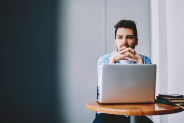 Ciddi odaklı, elleri bağlı genç bir adam, gündelik giysiler içinde modern dizüstü bilgisayarla çalışma masasında oturuyor ve gözlerini başka yöne çeviriyor.