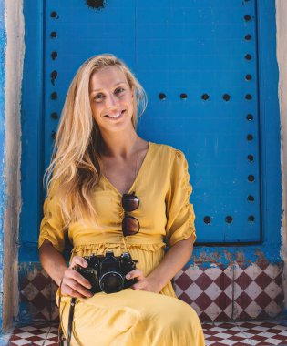 Fas 'ta seyahat sırasında yaşlı bir binanın kapısında oturmuş kameraya bakıp gülümseyen mutlu sarışın kadın.