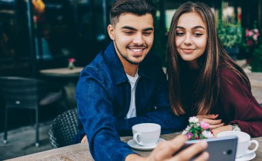 Kafe terasında masada oturan cep telefonu kamerası selfie için poz neşeli erkek ve kadın genç hipsters, kahve ile tarih boyunca cep telefonu üzerinden fotoğraf yapma çift birlikte zaman keyfini çıkarın