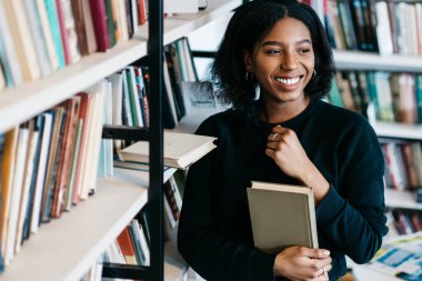 Afro-amerikan neşeli bayan, günlük giysiler içinde kitap alıyor ve kütüphanede kitap rafı başında dikilip eğleniyor.