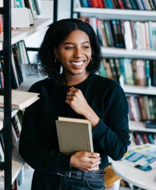 Afro-amerikan neşeli bayan, günlük giysiler içinde kitap alıyor ve kütüphanede kitap rafı başında dikilip eğleniyor.