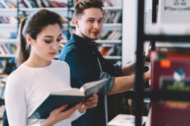 Genç, zeki, konsantre olmuş, günlük giysiler içinde kitap seçen ve okuyan öğrenciler, şehir kütüphanesinde kitaplarla karşı raflarda duruyorlar.
