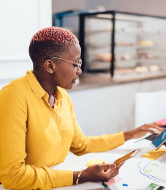 Kafeteryadaki iş görüşmesi sırasında elinde cep telefonuyla oturan ve mahsul meslektaşının akıllı telefonuna bakan modern Afrikalı Amerikalı kadın girişimcinin yan görüntüsü