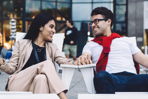 Joyful multiethnic couple holding hands with affection and talking while sitting on wooden chairs next to shopping bags on city street looking at each other