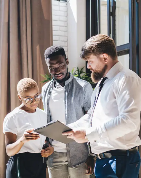 Side view of bearded male employee in formal wear holding clipboard discussing deal details with black man and woman standing at new home