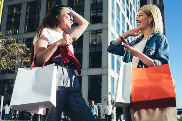 From below laughing cheerful stylish girlfriends with shopping bags standing on street during conversation after buying clothes together looking at each other