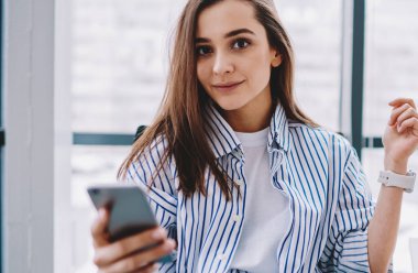 Güzel, siyah saçlı, şık giyinmiş genç bir bayan öğrenci masada otururken cep telefonu kullanıyor ve ofisteki kameraya bakıyor.