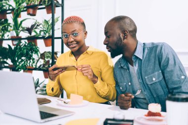 Genç Afrikalı Amerikalı bir kadın kafeteryada erkek meslektaşıyla çalışırken sosyal medyayı paylaştığı için akıllı telefonlu bir çantayla fotoğraf çekiyor.