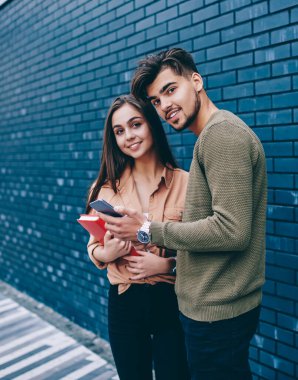 Genç erkek ve kız öğrencilerin birlikte akıllı telefon kullanarak ağlardan haber kontrol portresi, çift 4g bağlantısı kullanarak cep telefonu kamera tarama web sayfasına bakarak duvara yakın duran