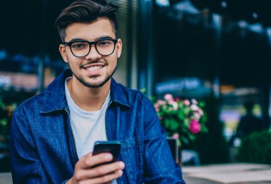 Gözlüklü sakallı hippi adamın portresi sohbet etmek ve sohbet etmek için cep telefonu kullanıyor. Kafenin içinde boş zaman geçiriyor. Neşeli milenyum erkeği kameraya bakıyor.