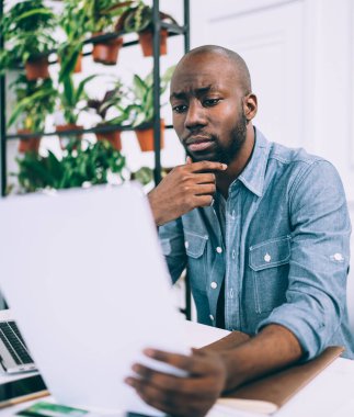 Ciddi bir Afro-Amerikan erkeğine odaklanmış günlük giysiler içinde çene ovuyor belgelerle çalışıyor ve dizüstü bilgisayarla sörf yapıyor yaratıcı stüdyoda masada oturuyor arka planda saksılarda çiçeklerle oturuyor.