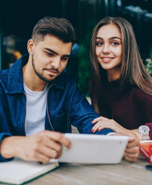 Pozitif genç kadın kamera bakarken erkek arkadaşı taşınabilir pc üzerinden online oyun oynamaya konsantre, erkek ve kadın hipsters video izlemek için cihaz kullanarak kafe terasında oturan